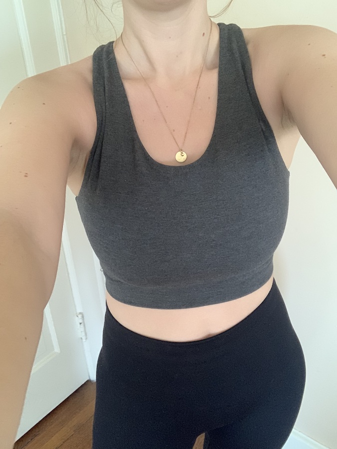 A person wearing a dark gray sports bra and black leggings, standing indoors, with a gold necklace visible.