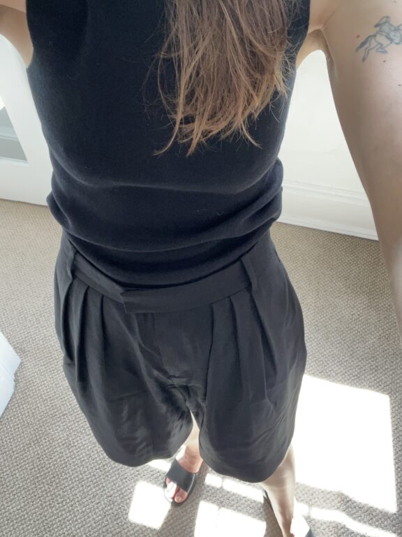 Person wearing a black sleeveless top, pleated black shorts, and black sandals standing on a carpeted floor in bright natural light.