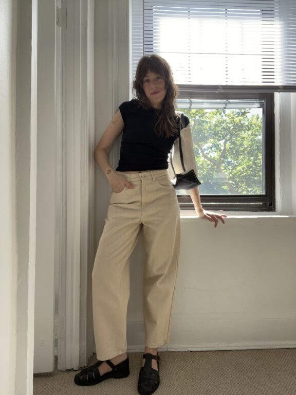 A woman in a black shirt and beige pants stands by a window with sunlight streaming in, one arm resting on the windowsill.