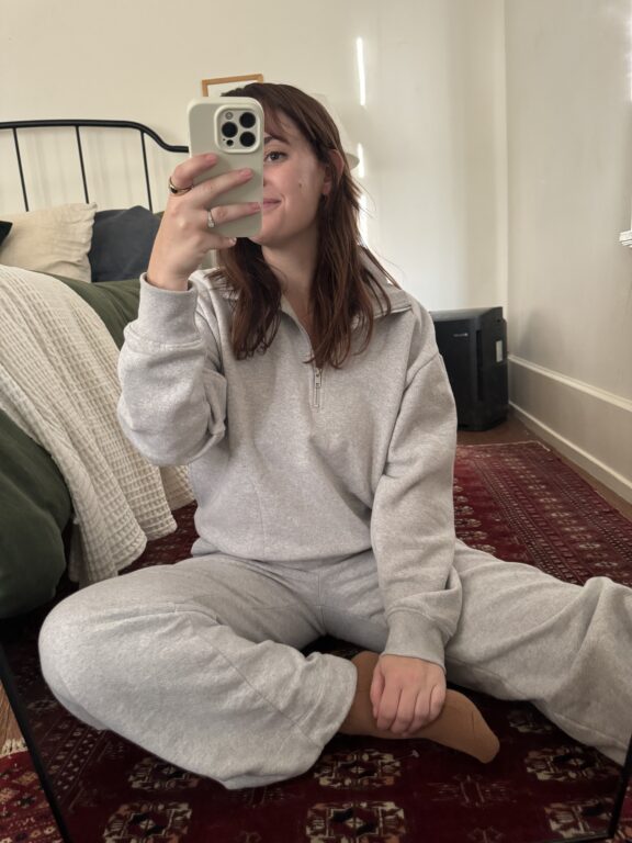 A person in a gray sweatsuit sits cross-legged on a red patterned rug, taking a mirror selfie in a bedroom with neutral decor.