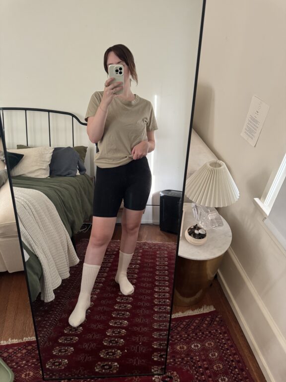 Person taking a mirror selfie in a bedroom, wearing a beige t-shirt, black shorts, white socks, and holding a phone. Bed, side table, and lamp are visible in the background.