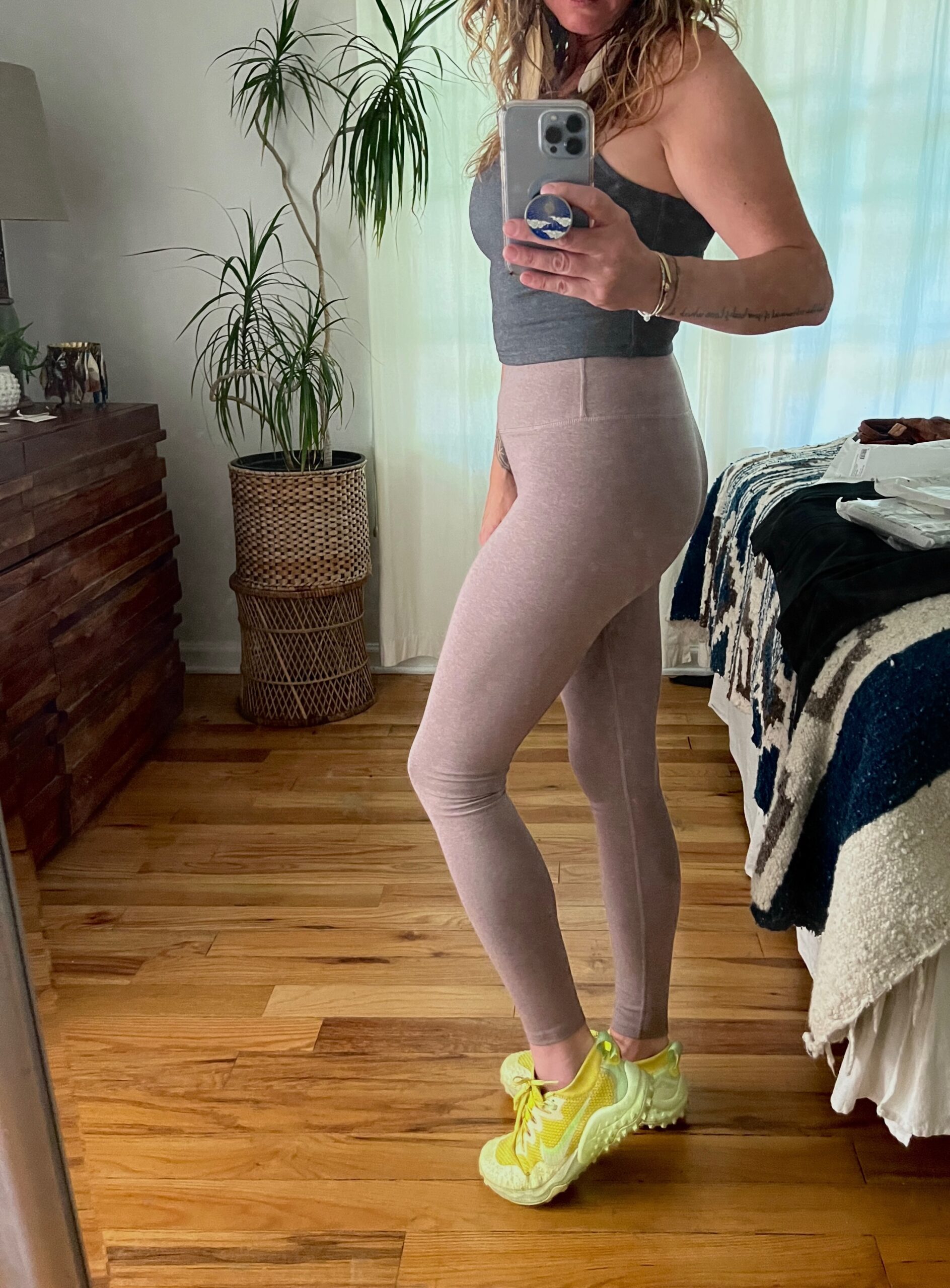Person taking a mirror selfie in a bedroom, wearing a gray tank top, light pink leggings, and yellow sneakers. Houseplants and furniture are visible in the background.