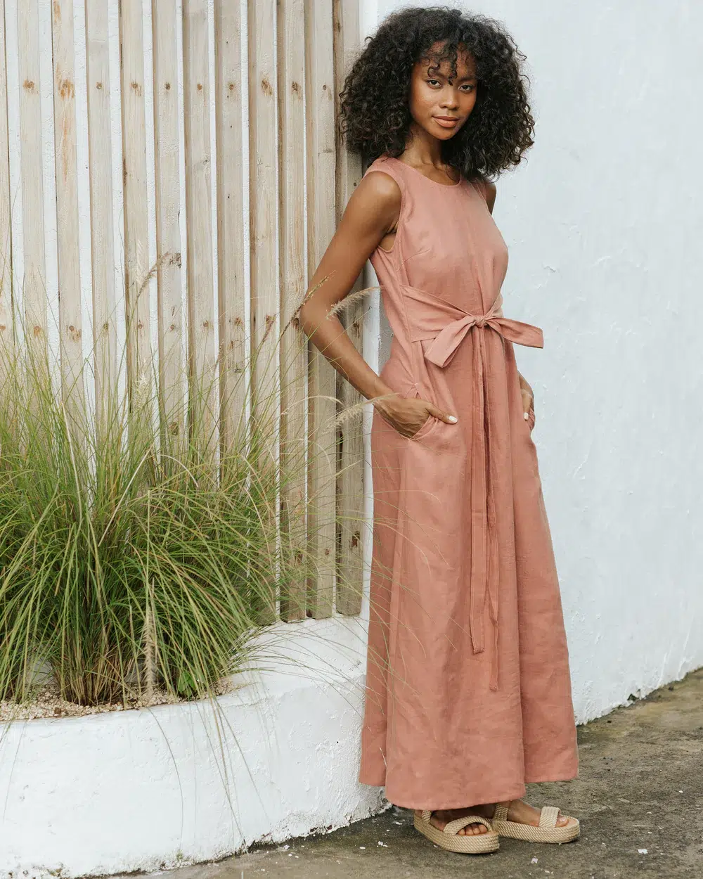 Woman standing outdoors wearing a sleeveless, ankle-length, pinkish-brown dress with a waist tie and sandals, next to a wooden fence and tall grass.