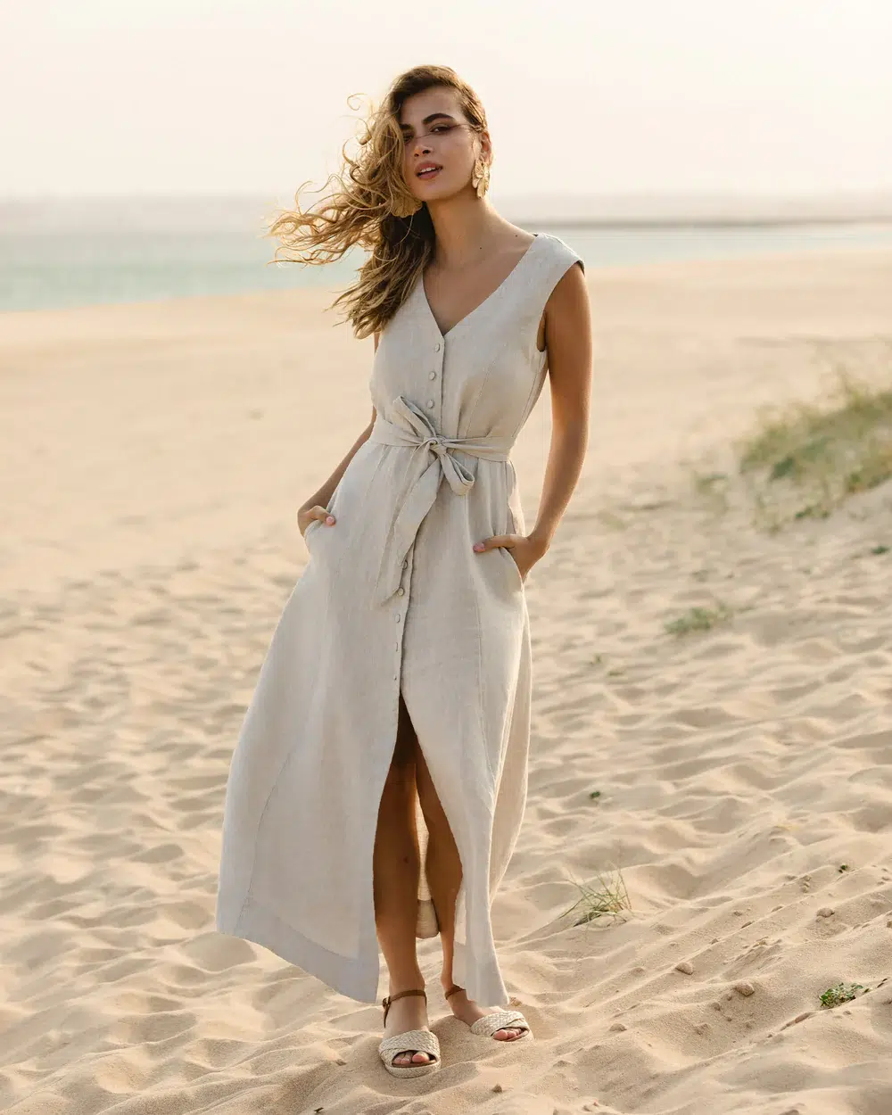Woman stands on a sandy beach wearing a sleeveless, button-down beige dress with a waist tie and sandals. The ocean and sky are visible in the background.