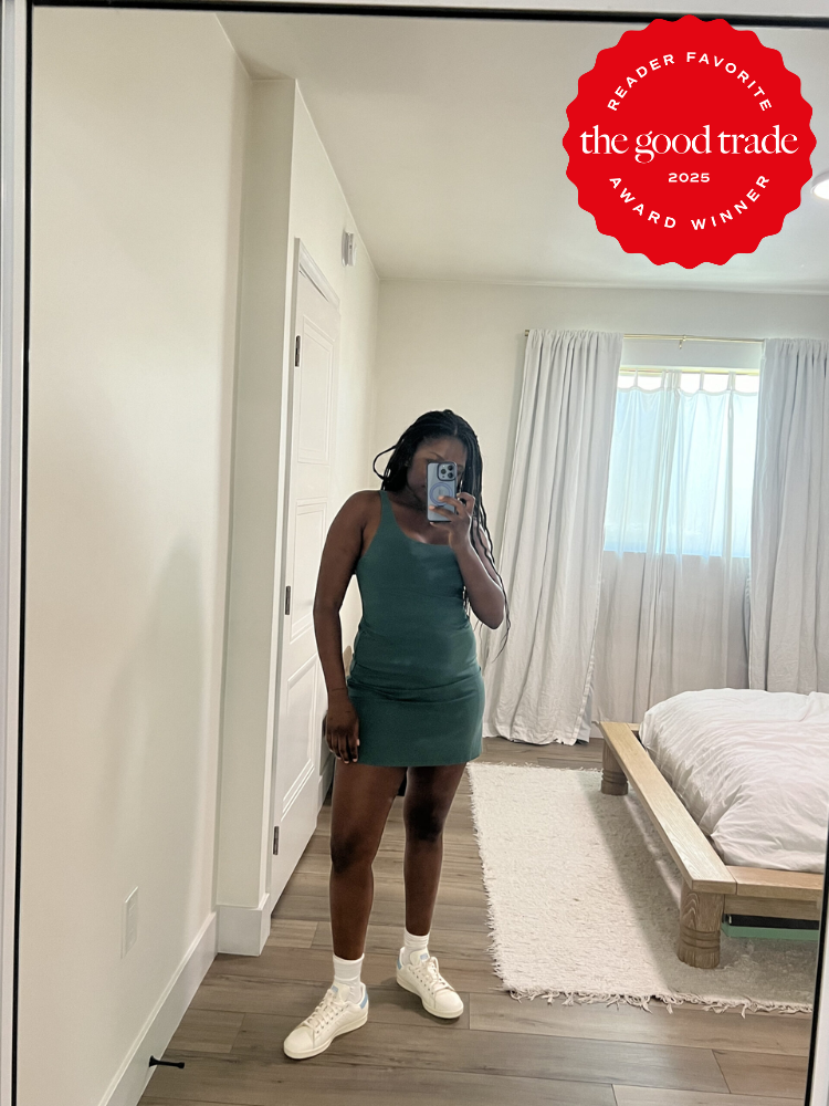 A person stands in front of a mirror taking a selfie, wearing a green dress and white shoes in a bedroom. A "FashionGroomSpot 2025 Award Winner" badge is visible in the top right corner.