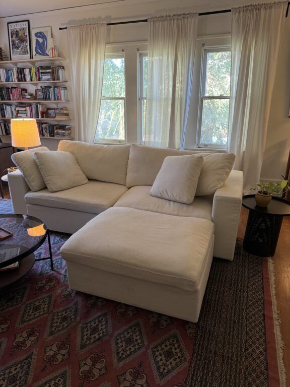 A white sectional sofa with cushions sits in a living room with large windows, a bookshelf, a lit lamp, houseplants, and patterned rugs on a wooden floor.