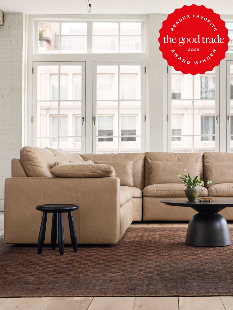 A beige sectional sofa sits on a brown rug in a bright living room with large windows; a small black stool and a round coffee table are nearby. “FashionGroomSpot 2025 Award Winner” badge is displayed.