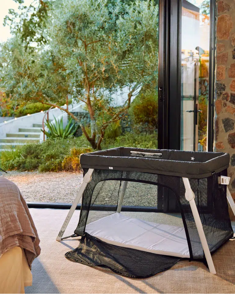 A black and white portable crib is set up indoors near a large glass door, with a garden visible outside.