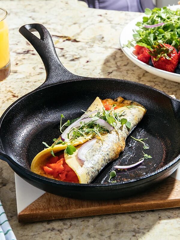A rolled omelette with vegetables and greens sits in a cast iron skillet on a kitchen counter beside a glass of orange juice and a salad.