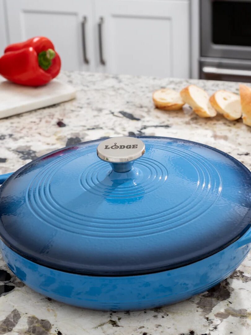 A blue Lodge cast iron casserole dish with handles sits on a kitchen counter near sliced bread and vegetables.